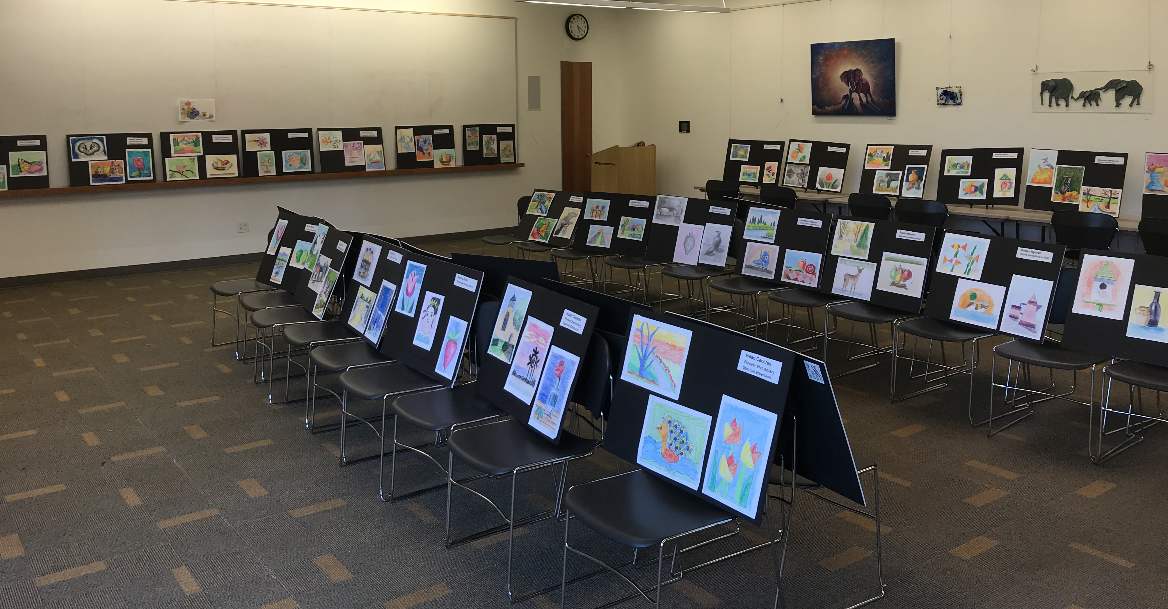 Rows of student paintings displayed on easels and chairs at a student art exhibition